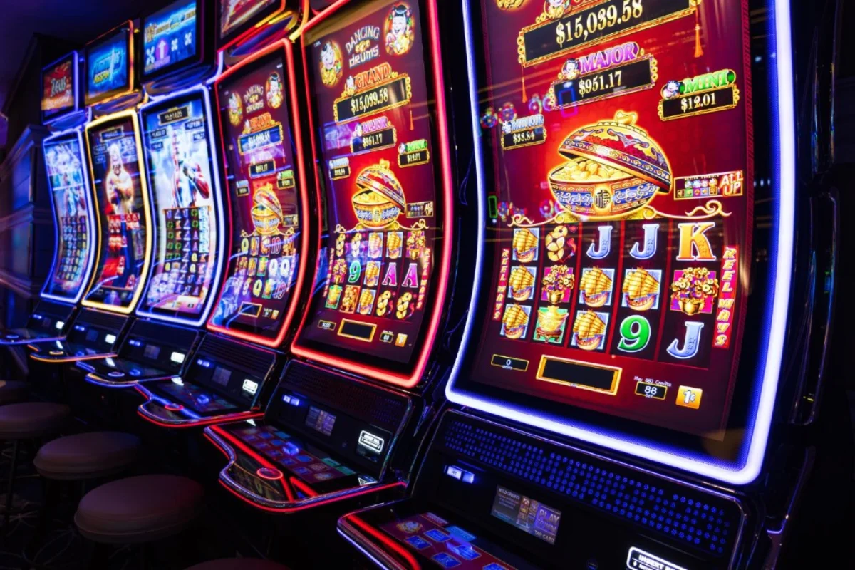 Row of brightly lit modern slot machines on a dim casino floor with colourful Asian-themed reel displays